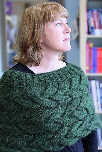 A woman wears a green cabled shrug that wraps around the upper portion of her body like a big cowl.