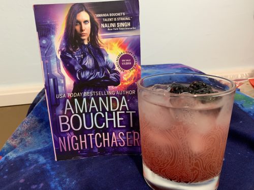 A light purple drink in a tumbler glass next to a copy of Nightchaser. Both are displayed on a purple and blue scarf.