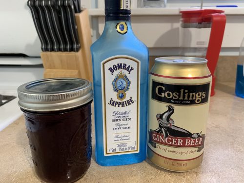 A jar of blackberry shrub syrup, a can of ginger beer, and a bottle of gin on a kitchen counter.