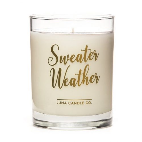 A white candle in a glass contained. Sweater Weather is printed in gold lettering.