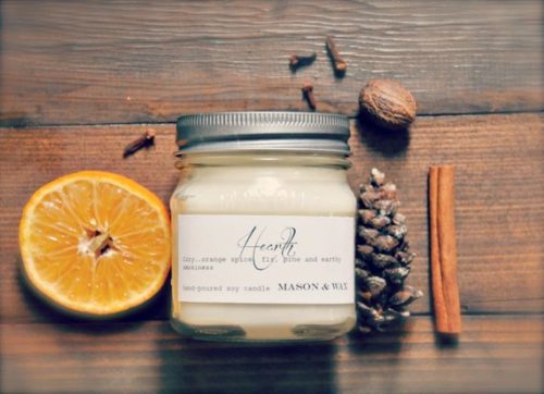 A white candle in a square jar. It's resting on a wooden table with a cinnamon stick, chestnuts, and oranges.