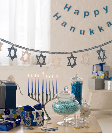 A garland of the Star of David crocheted in white and blue is hung over a table set with a menorah, gifts and candies.