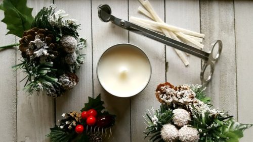 A white candle in a metal tin surrounded by pine needles and holly berries.