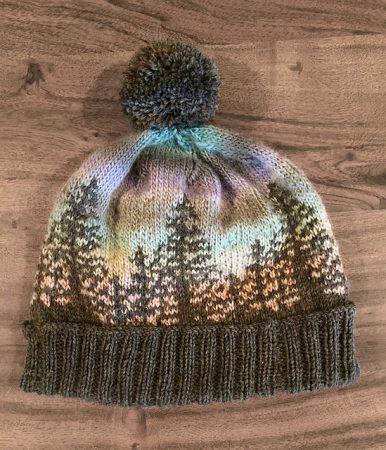 A beanie style hat that has a brown brim with the outline of fir trees in the same brown. The background is made with an ombre yarn so it looks like the trees are silhouetted against a sunset.
