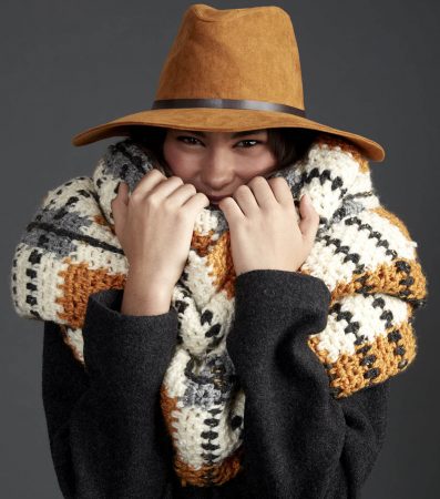 A woman in a super cute hat is all snuggled up in a giant tartan scarf. The main color is cream with contrasting stripes of brown, black and gray