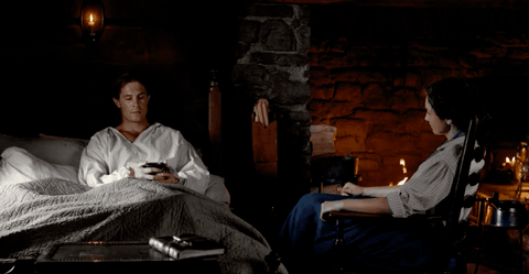 John, in bed, holding a cup of tea, and Claire sitting in the rocking chair next to the bed.