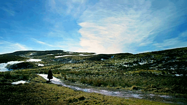 A still of Brianna walking down a road with the sun and the green grass and snow and a huge blue sky