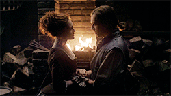 Brianna and Roger kneeling in front of the fire.