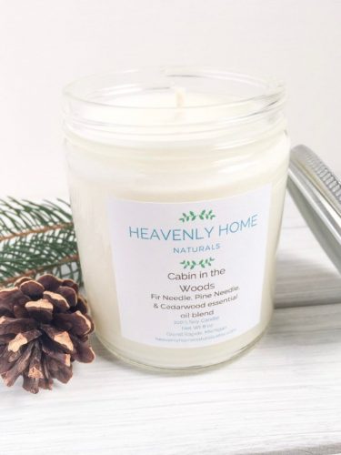A bright white candle with a label with light blue lettering. It's displayed next to some pinecones and pine needles.