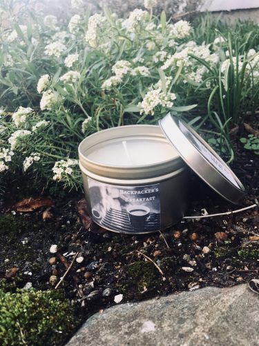 A candle in a metal tin. It's outside in the dirt next to a rock and some wildflowers.
