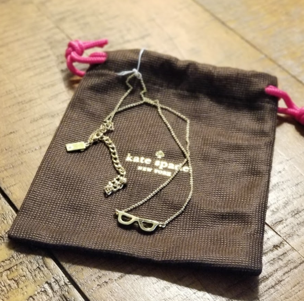 Kate Spade gold plated necklace with mini eyeglasses charm sitting on a brown fabric bag printed with the kate spade logo 