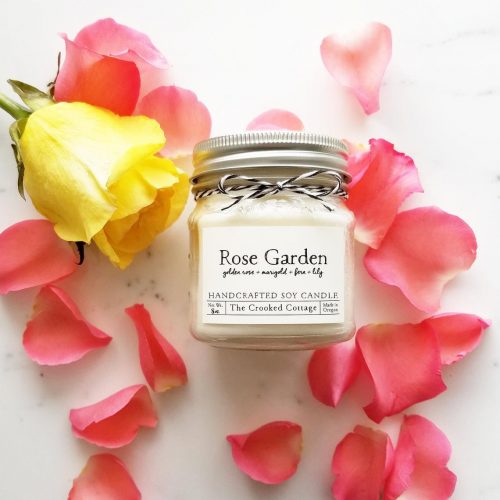 A candle in a square jar amidst some light pick rose petals.