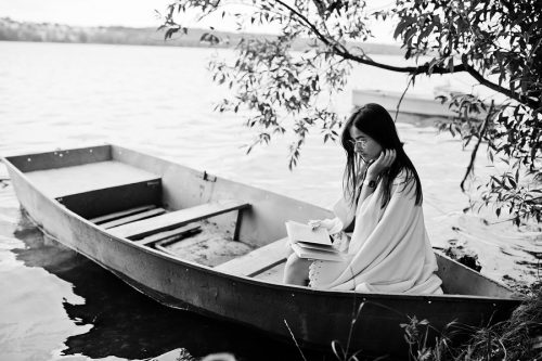Portrait of an attractive woman wearing black polka dots dress white shawl and glasses reading a book in a boat on a lake. Black and white photo.