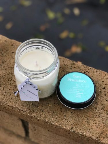 A white candle in a glass jar resting on a stone ledge. The lid is black with a teal label.