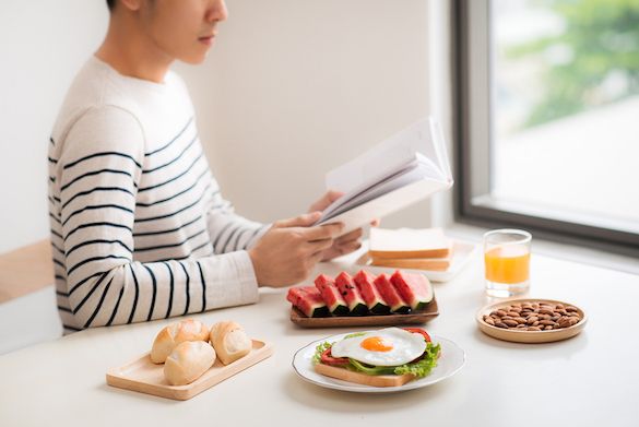 A man eating breakfast and reading book at home in morning. hes' got fruit a fried egg some buns and a glass of juice but zero utensils 