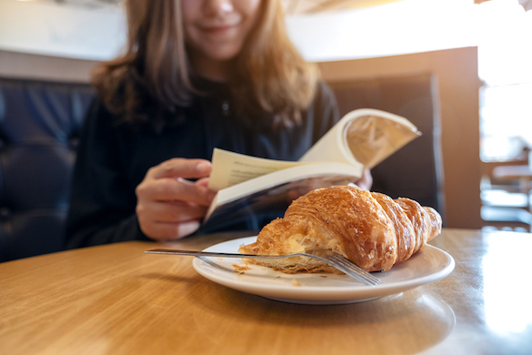 Closeup image of a woman holding and reading a book with a piece of croissant in a plate on wooden table again with the fork who uses a fork?