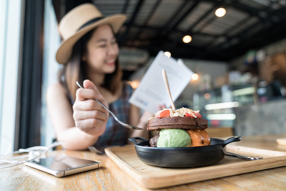 Happy Asian woman wearing dress with brown hat sitting and eating ice cream with chocolate waffles ontop with almond and fruit serve on wooden plate in dessert caf while reading a book.