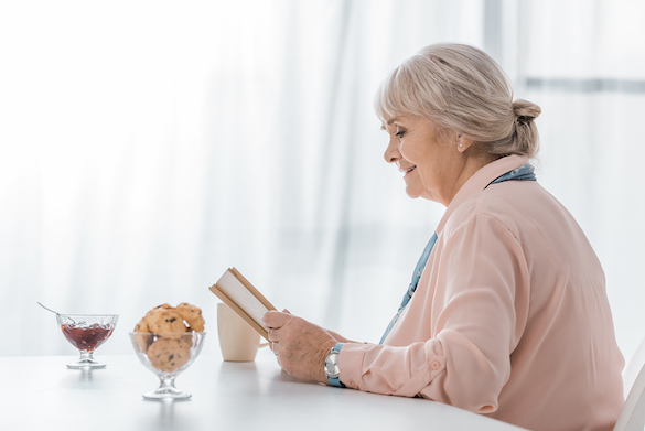senior woman sitting at table and reading book in nursing home wearing a pink cardigan with a glass full of puffy cookies and a jar of cherries and a drink on the table with her 