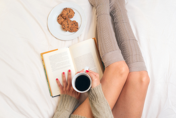 Woman eating breakfast in bed while reading a book . She is photographed from above wearing knee socks and holding her book beside her knees and it all looks very uncomfortable 