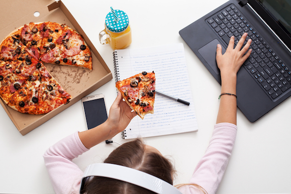 Young teenager girl working on a project while eating pizza at the desk - using her laptop, smartphone and exercise book, top view
