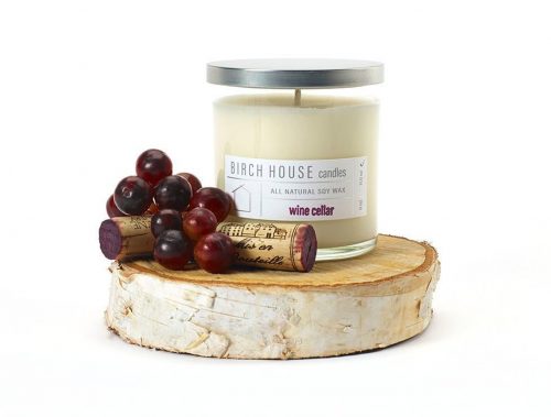 A white candle in a clear jar. The jar rests on a wooden birch platform and is next to a pile of wine corks and grapes.
