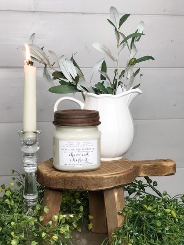 A white candle in a square jar. It's on a wooden stool next to a lit candlestick and a white porcelain pitcher filled with greenery.