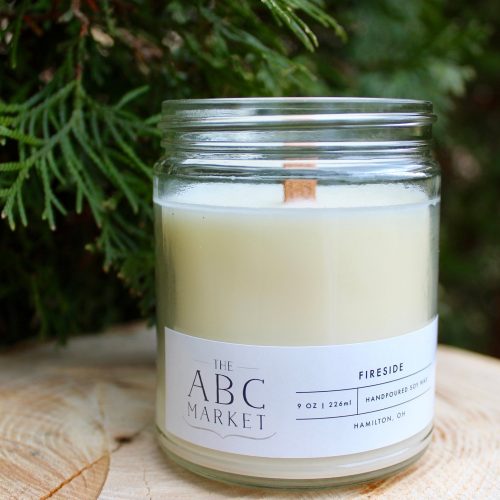 A white candle in a glass jar. It's sitting on a tree stump next to a fir tree.