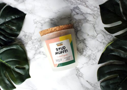 A candle in a frosted glass jar with a cork lid. The label is white, peach, yellow, and green. So colorful and fun! It's also situated in front of a marble background.