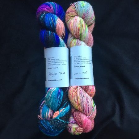 Two skeins of yarn against a dark blue background. The skein on the left is a deep blue with bright purple and orange varigations. The skein on the right is a neon pink with neon yellow, green and orange varigations.