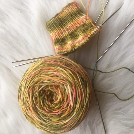 A cake of yarn and the cuff of a sock against a white background. The yarn is a pale green with pooling in light pink and cream, like the colors of a rainbow trout