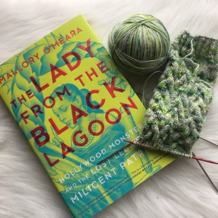 A copy of the book The Lady from the Black Lagoon and a cabled sock leg in varying shades of green. A ball of the yarn sits in between them.