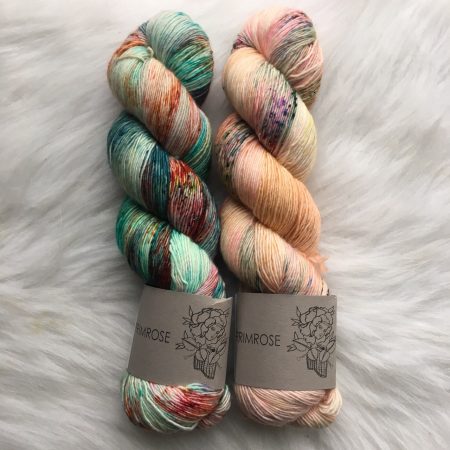 2 skeins of yarn side by side on a white background. The skein on the left is green with red and peach speckling. The skein on the right is a peachy-pink with some red and blue varigation.