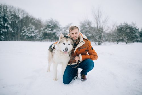 Young man sits in the snow next to a dog, an Alaskan Malamute.