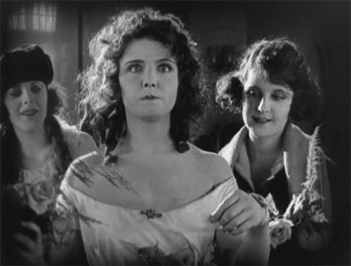 A black and white film clip of a woman with brown curly hair in what looks like a white bridal dress fainting into the arms of her friends who catch her, thank goodness.