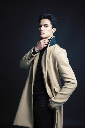 Handsome Asian man posing in studio on black background. He's wearing a long, tan coat with a black collar.
