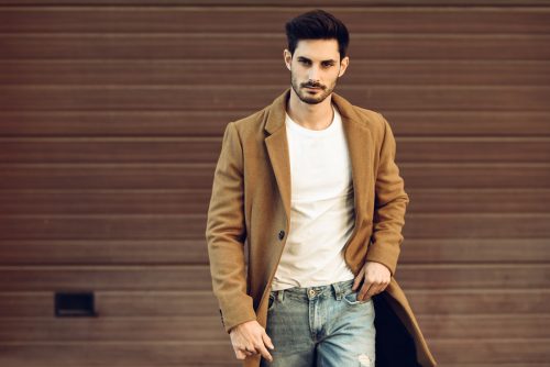 Young man in front of a wooden wall. He's got some lovely scruff. He's wearing a tan coat with jeans and a white t-shirt.