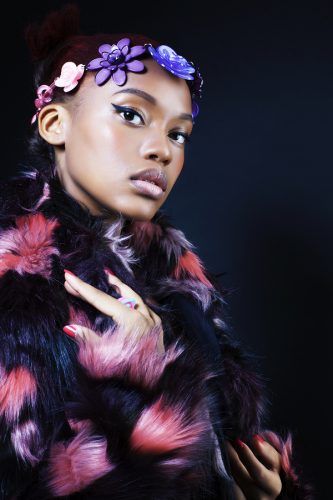 Young pretty African American woman in a black furry coat with pink and purple spots. She has winged eyeliner on and a flower headband.