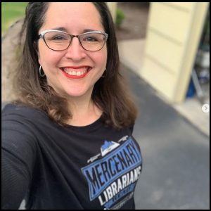 AnaCoqui smiling wearing a fabulous lipstick color wearing a MERCENARY LIBRARIANS READ shirt