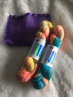 two skeins of teal and gold and coral yarn and a purple yarn tote against a fluffy background
