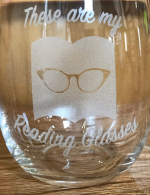 A stemless wineglass with THESE ARE MY READING GLASSES etched in the side above an image of retro glasses a top an open book