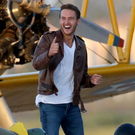 Peter wears an aviator jacket and gives a thumbs up in front of a plane.