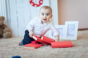 A very surprised looking baby on a beige carpet, crawling on top of some red covered books with a wide eyed exhilirated epxression