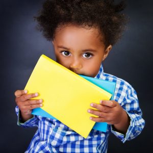 Closeup portrait of cute little shy American schoolboy with textbooks in hands over blackboard background, ready to study in elementary school