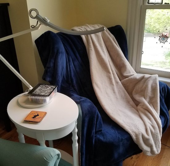 My favorite reading chair by a window with the lamp, my kindle, a bunch of fluffy blankets and my cross stitching box on a side table the lamp is long and thin with a curved head and a white base and body 