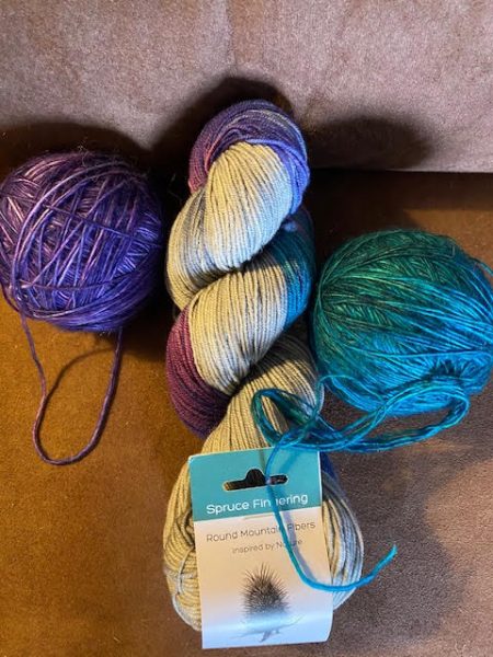 Purple and teal balls of yarn sit on either side of a skein of light gray shot through with some of those colors