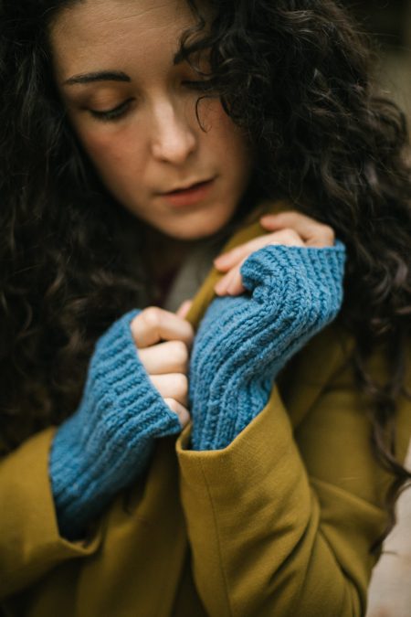 A white woman with long curly brown hair models a pair of blue ribbed fingerless gloves