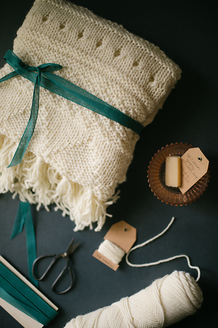 A cream colored baby blanket is folded and wrapped with a ribbon. Some yarn and scissors lay next to it