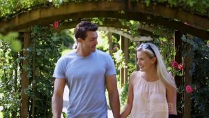 Art and a young blonde woman walk through a flower arch