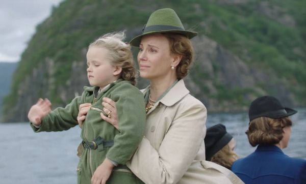Martha in a green hat holding up her son in a green coat to wave goodbye on a windy boat clearly very upset 