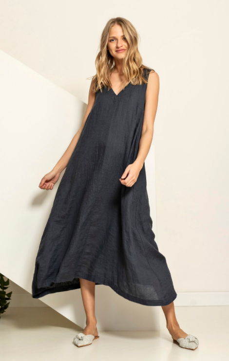 A blonde model wearing a loose fitting blue linen shift dress that billows out at the hem and comes to mid-calf